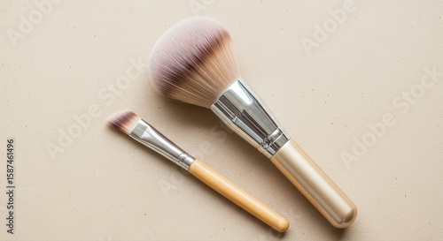 Two makeup brushes, one powder brush and one smaller eyeshadow brush, lie on a beige surface