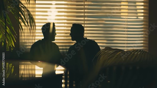 Two silhouetted figures converse near a window with blinds