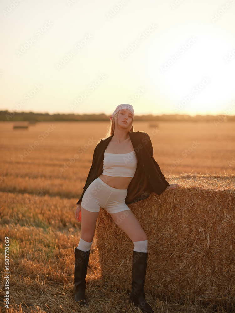 Young stylish woman in white crop top, shorts, jacket and bandana, posing near hay bale on wheat field at sunset. Young stylish woman in white crop top, shorts, jacket and bandana, posing near hay bale on wheat field at sunset.