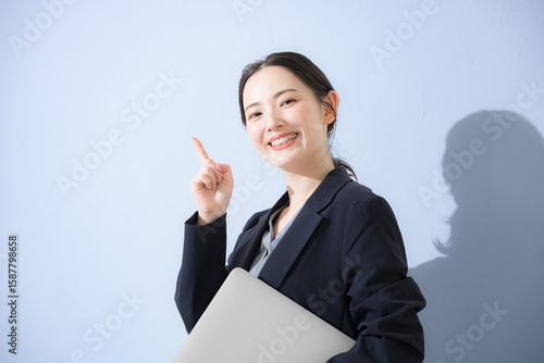 Photography パソコン持ってスーツを着た立つポイントなどの指差しポーズの若いかわいい女性のビジネス上半身　水色背景の営業などリクルートのイメージ