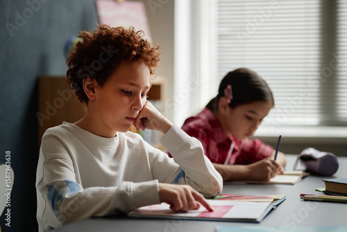Caucasian boy reading book at desk while teenage girl with dark hair writing in notebook in classroom setting, both focused on studying, natural light coming through window