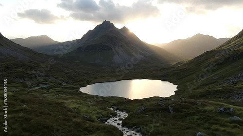 Lake nestled amidst rolling hills and mountains illuminated by sunlight filtering through the clouds