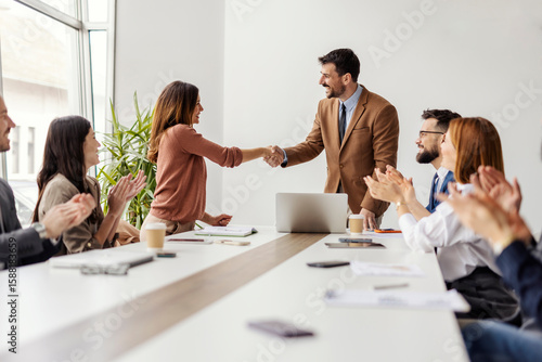 Happy businesswoman shaking hands with ceo while leadership team sitting at conference table and applauding.