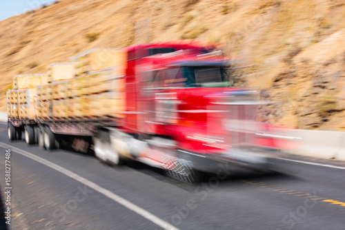 Semi-truck with a red cab with a load of timber, driving fast on a mountain road.