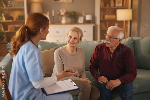 A home health aide, wearing scrubs and holding a clipboard, speaks to a senior couple sitting on a couch in their living room, discussing details of their care and needs.
