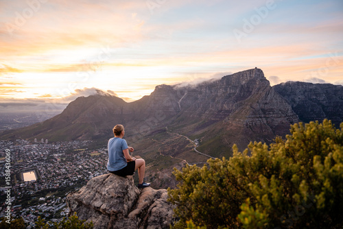 Eine Person sitzt auf einem Felsen und blickt über Kapstadt und den Tafelberg bei Dämmerung