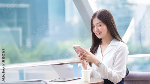 Wide 16:9 shot of young Asian businesswoman working outside office at coffee shop, using smartphone for remote communication and online business in urban city