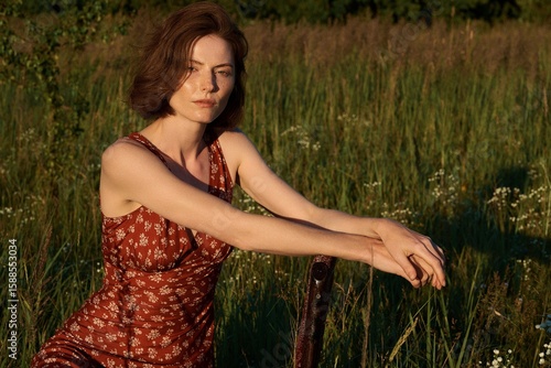 Tableau sur toile A woman with auburn hair and freckles sits in a sunlit field, looking thoughtfully at the camera
