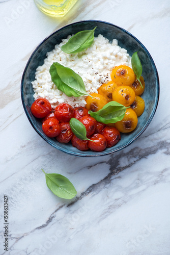 Bowl with cottage cheese, roasted tomatoes and green basil on a white stone background, vertical shot with space, above view