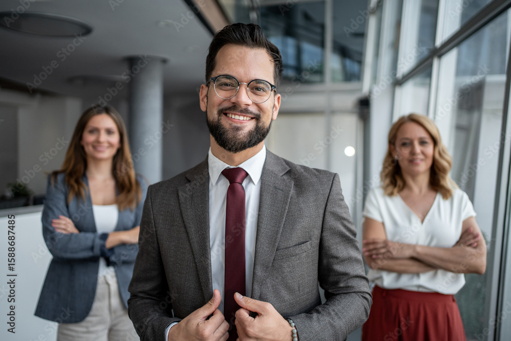 Smiling businessman adjusting suit with coworkers in background in office Smiling businessman adjusting suit with coworkers in background in office