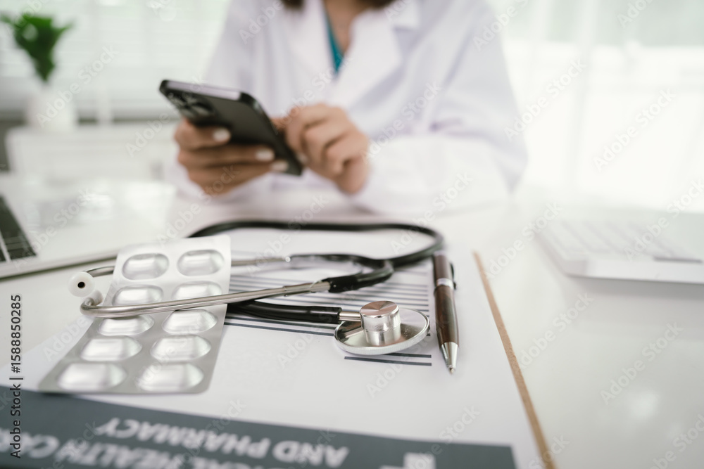 Fototapeta premium A doctor in a white coat uses a smartphone at a desk with a stethoscope, pills, medical documents, and a pen, focusing on healthcare communication and patient care.