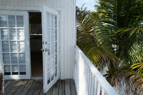 White French doors with room number 13 open onto a tropical balcony with wooden decking and lush palm leaves. Bright, airy, and inviting coastal ambiance.