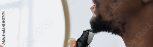 Cropped view of african american man with vitiligo shaving beard with electric razor in bathroom, banner