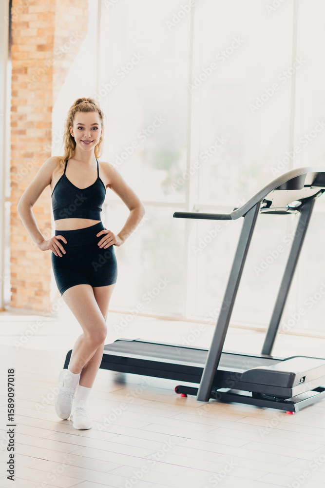 Fototapeta premium Active athletic woman in activewear standing confidently near a treadmill in a modern well-lit gym interior space