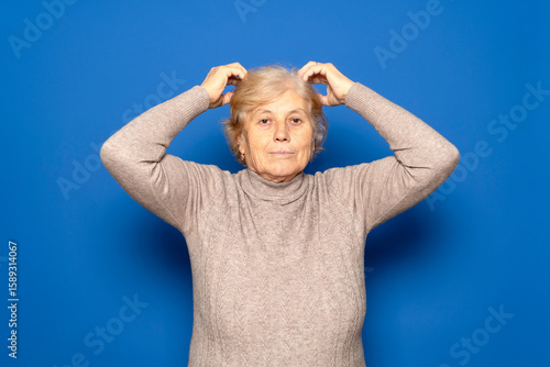 Caucasian woman in her 70s wearing a dark beige sweater scratching her head worriedly trying to remember something important, isolated over blue background.