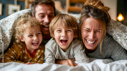Happy family of four laughs together under a blanket in bed, creating a joyful and loving family moment.