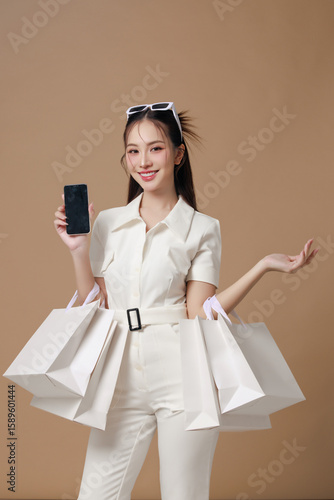 Young Asian woman in stylish outfit holding shopping bags and smartphone in studio against beige background