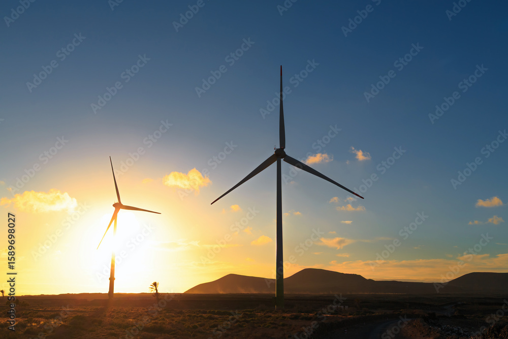 Obraz premium Two wind turbines stand tall against a vibrant sunset, with mountains and clouds in the background creating a peaceful atmosphere.