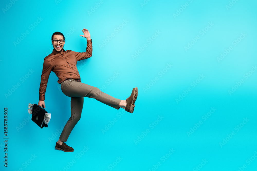 Fototapeta premium Dynamic young businessman jumping joyfully holding a black briefcase full of money on a bright teal background