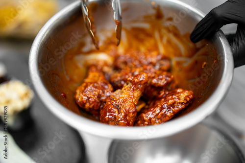 Close-up of crispy chicken wings coated in glossy sauce, tossed in a stainless steel bowl with tongs, showcasing appetizing texture and rich color, ideal for food photography.