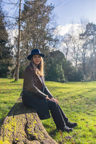 Smiling woman in dark coat and blue hat sitting on a mossy log in sunny park