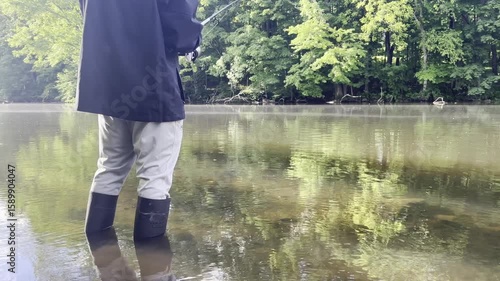 Angler dressed in full fishing gear stands near the water’s edge, raising his fishing pole at a high angle above the lake as the line stretches into the water.