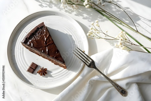 Slice of Rich Chocolate Cake with Star Anise and Chocolate Pieces on White Plate with Fork and Flowers
