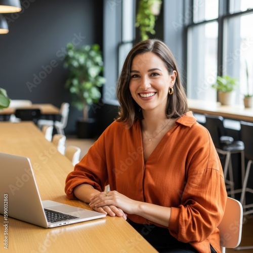 Smiling woman in orange shirt working on laptop in modern office setting