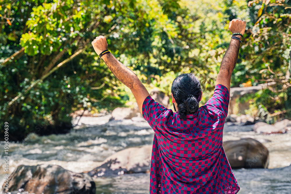 Obraz premium Man in plaid shirt with tied hair raising arms beside river in lush Colombian forest at countryside hotel in Santuario, Risaralda, with space for text