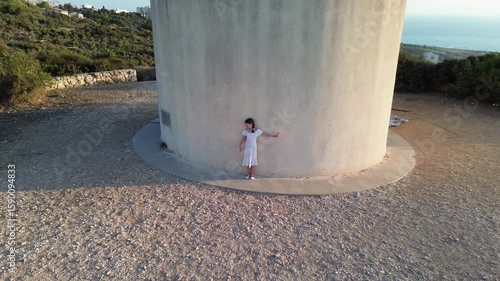 Aerial view from above of dancing young  beautiful blonde girl in white dress near the small church on the mountain Carmel near Haifa, Israel outdoors.  Shooting from the drone that flying away