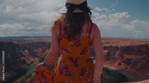 Rear view of a woman in a floral dress and sunhat overlooking the vast canyon landscape and the winding Colorado River at Horseshoe Bend.
