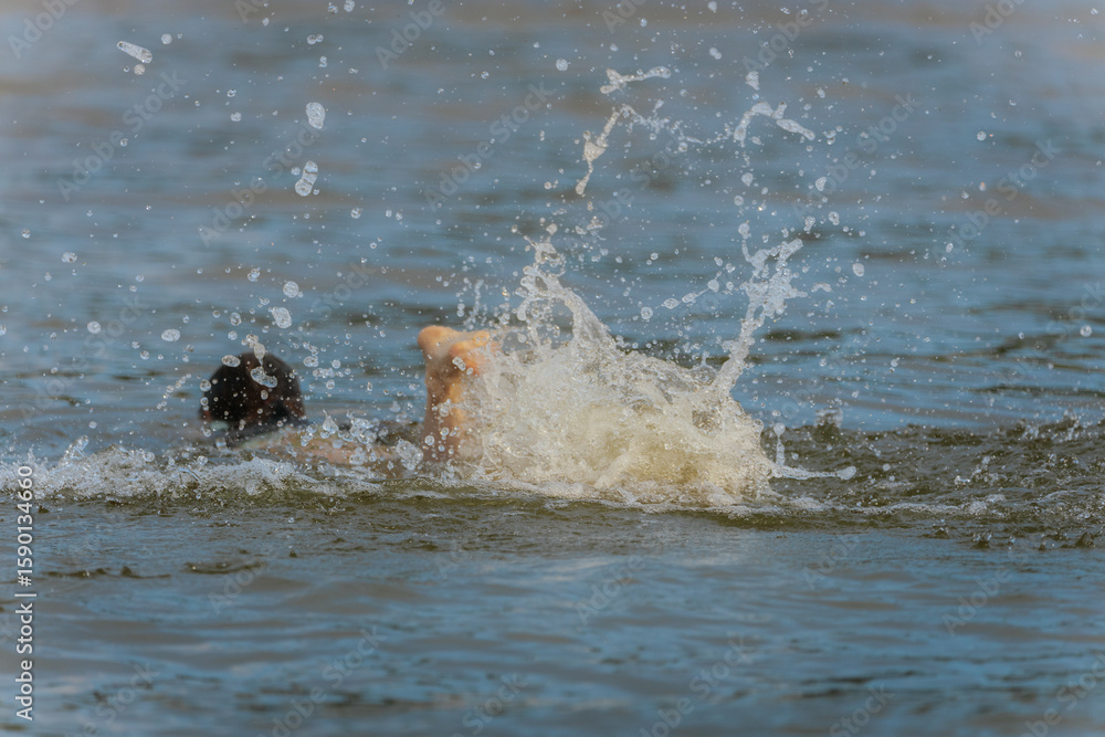 Obraz premium A smiling young boy stands shirtless near the water, flashing a peace sign with his fingers on a bright day at the beach.
