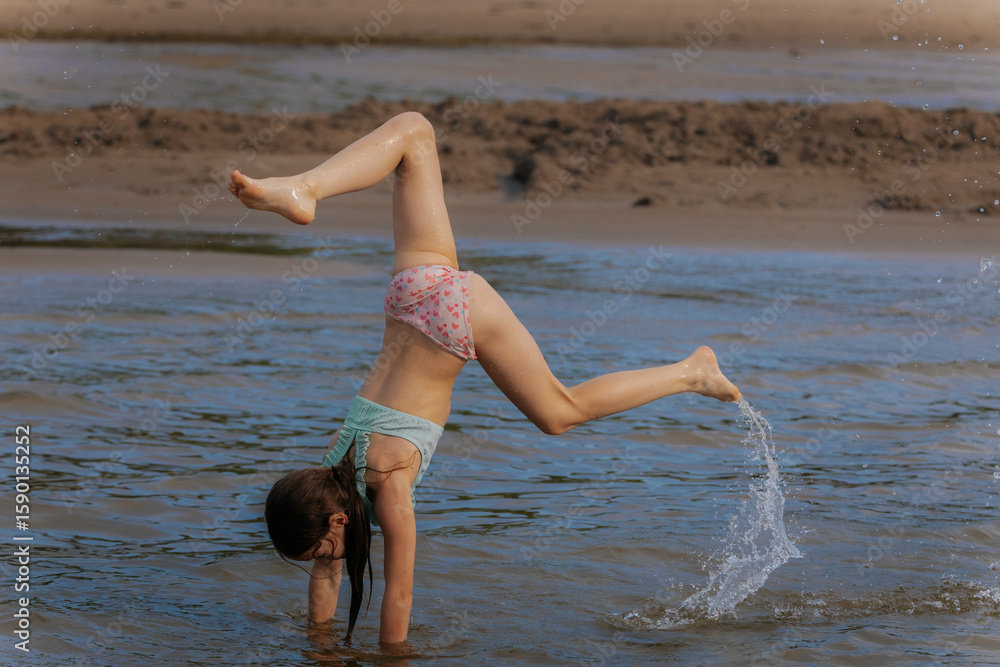 Naklejka premium A young girl performs a handstand in shallow water at the beach, showcasing strength and playfulness during a sunny day.