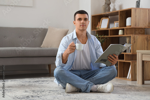 Handsome young man with cup of coffee reading magazine while sitting on floor...