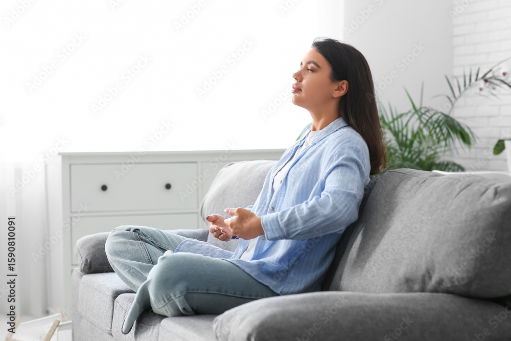 Fototapeta premium Beautiful young woman doing breath practice and meditating on sofa at home