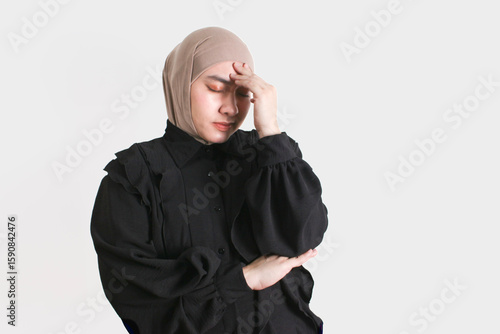 Portrait of a young woman wearing hijab and black blouse, holding her head in pain or stress, eyes closed. Expression of headache, anxiety, or mental exhaustion.