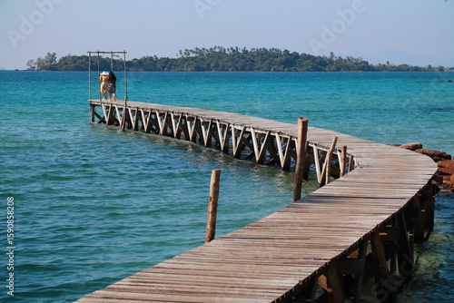 Canvas-taulu Curved wooden pier extending into turquoise waters with a distant of couple and island backdrop in Klong Mad fisherman village, Koh Kood, Thailand