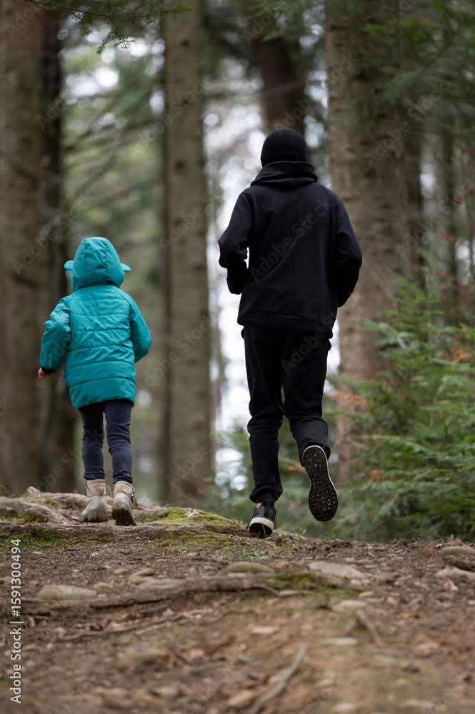 Fototapeta premium Joyful exploration on a serene forest trail during a chilly day as a young child and adult walk together amidst tall trees and a soft carpet of fallen leaves