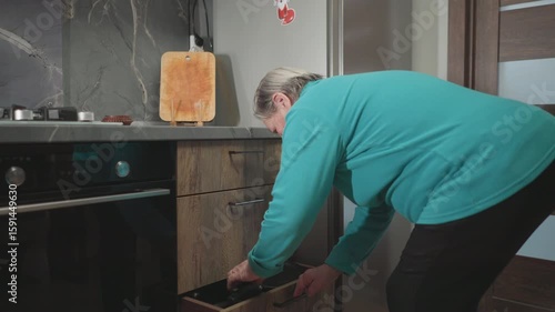 Elderly woman in green sweatshirt bending over to open kitchen drawer, searching for something. Modern kitchen with wooden accents and a marble countertop, capturing everyday routine in a home setting
