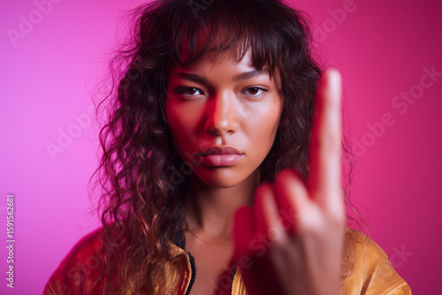 Confident young woman with curly hair gestures assertively against a vibrant pink backdrop while wearing a golden jacket, expressing self-assurance and determination