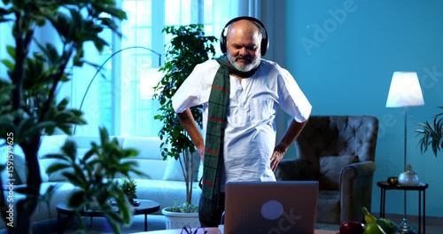 Indian senior man doing online exercise session using laptop in modern home interior, happy aged Asian male following virtual fitness routine indoors to stay healthy, active, and physically fit