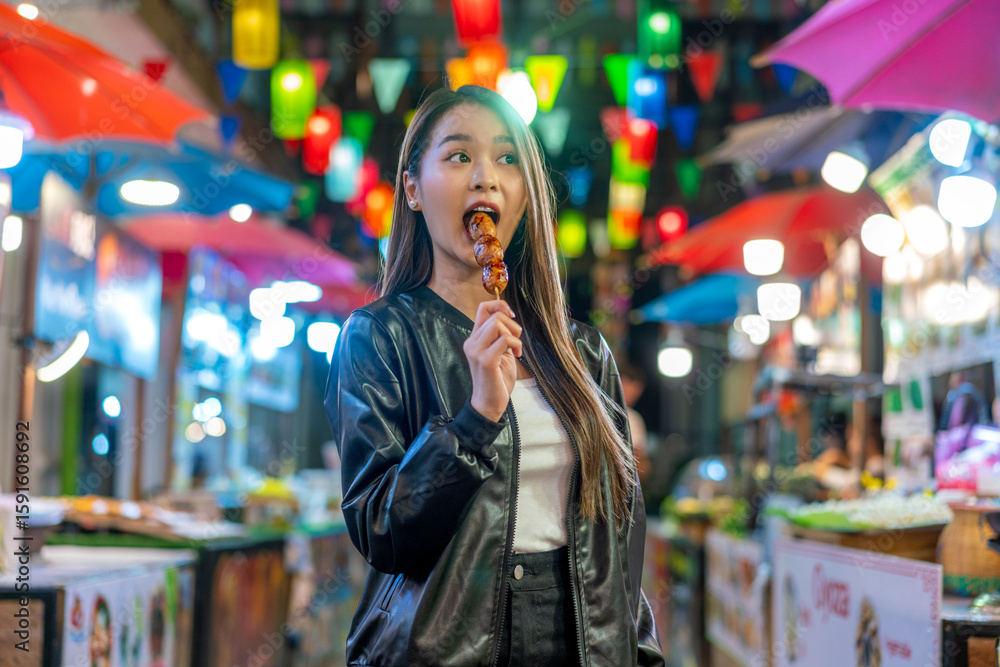 Fototapeta premium Asian woman enjoying street food at Chiang Mai Walking Street Night Bazaar. Young traveler exploring vibrant night market in Thailand.