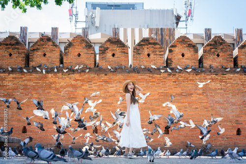 Asian tourist woman enjoying travel moments at Tha Phae Gate, Chiang Mai. Young woman in white dress surrounded by flying pigeons in historical city square.