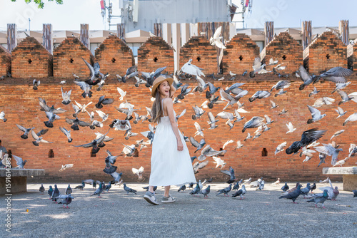 Asian tourist woman enjoying travel moments at Tha Phae Gate, Chiang Mai. Young woman in white dress surrounded by flying pigeons in historical city square.