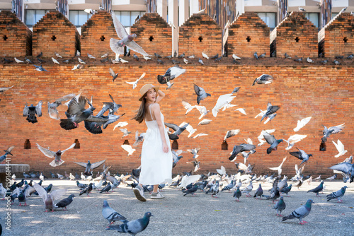 Asian tourist woman enjoying travel moments at Tha Phae Gate, Chiang Mai. Young woman in white dress surrounded by flying pigeons in historical city square.