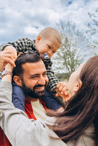 Close-up of a family,a toddler sits on dad shoulders, crying and reaching out to mom. She gently holds his hands, trying to comfort him. A touching moment of parenting and family connection