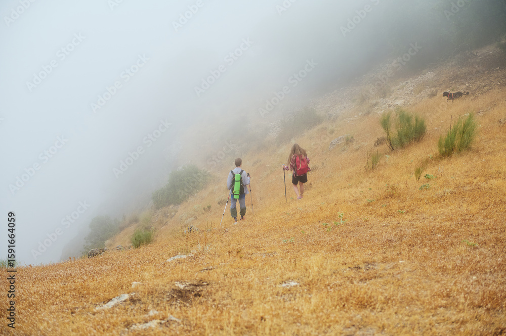 Obraz premium Couple Hiking on a Foggy Mountain Trail Through Dry Grassland and Mist
