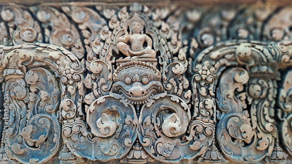 Obraz premium Intricately carved sandstone relief from Banteay Srei temple in Siem Reap province, Cambodia, depicting a guardian figure and Hindu deity in detailed floral and mythological patterns.