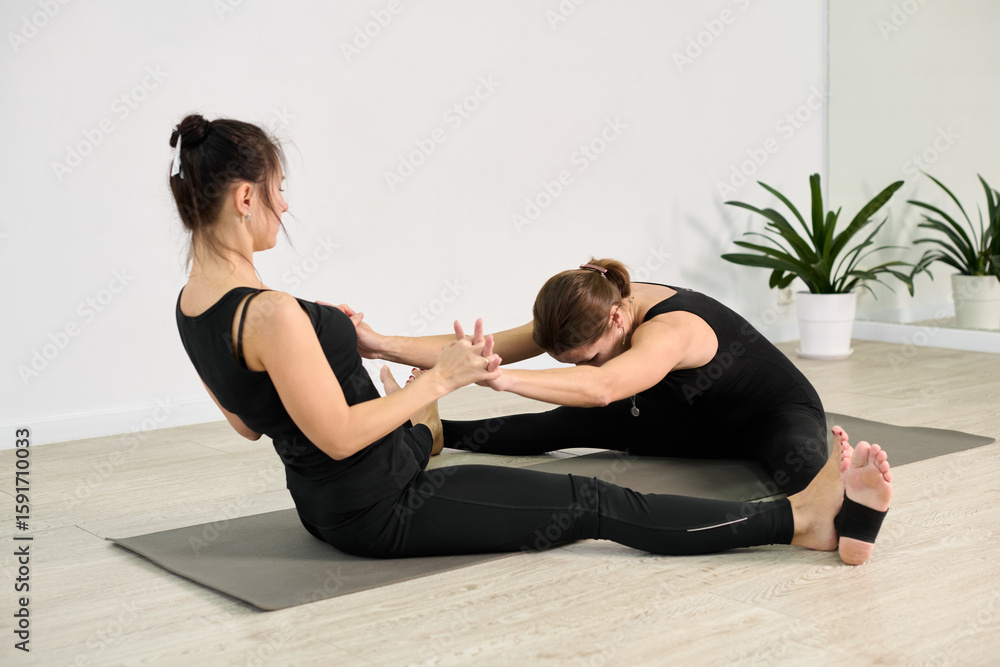 Obraz premium Two young women practice partner yoga in studio. They sit on mats, stretching forward in synchronized pose. Light walls and plants create calming ambiance