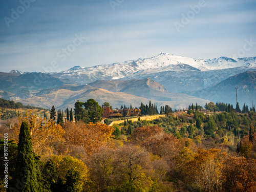 Stunning view of Sierra Nevada from Alhambra’s Alcazaba in Granada, Spain, with autumn foliage, historic stone walls, and snow-capped peaks under a clear, vibrant sky.